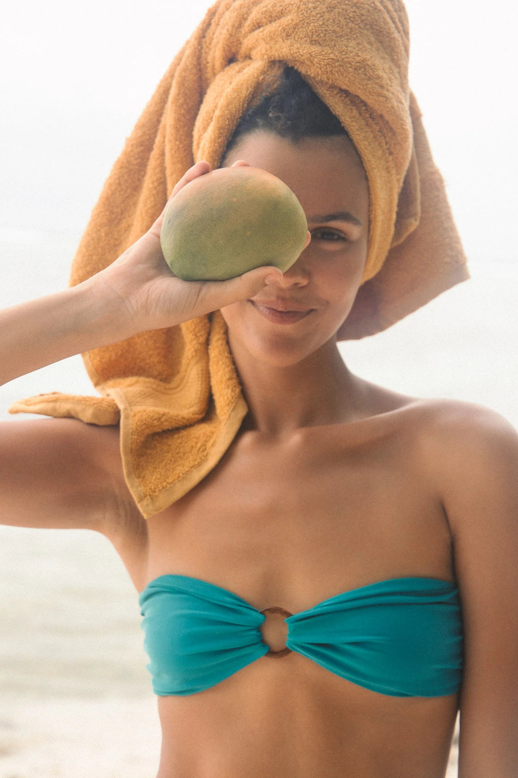 Woman in teal bikini with towel on head, holding a mango against face.